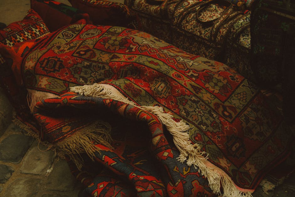 A close-up view of an intricately patterned, multicolored Persian rug with dominant red, navy, beige, and green tones, draped over a surface, revealing fringed edges and detailed geometric and floral motifs. The rug's surface appears clean and well-maintained, highlighting its vibrant colors and fine weaving. The setting suggests an indoor space with other upholstered furniture nearby, and the lighting emphasizes the texture and craftsmanship of the rug. This image showcases surface cleaning and maintenance as part of a professional domestic or commercial cleaning service offered by carpetcleanerswestkensington.co.uk, specifically under their deep cleaning and sanitisation offerings for rugs and carpets on Kensington High Street, W8.