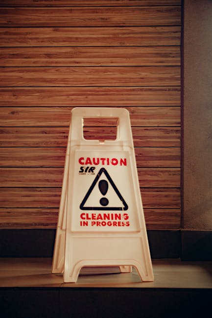 A white plastic 'Caution Cleaning in Progress' warning sign with black and red text and a triangular warning symbol, placed on a light wooden floor against a background of vertically aligned wooden paneling on the wall. The sign is positioned near the corner of a room, indicating ongoing cleaning activities by [COMPANY_NAME], part of their domestic cleaning and surface sanitisation services at the West Kensington location. The lighting is even, highlighting the contrast between the sign and the warm-toned wood surfaces, emphasizing a clean and safe environment.