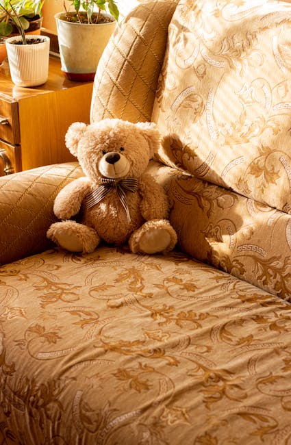 Close-up of an upholstered sofa in a residential living room, featuring a beige fabric with intricate floral patterns. The sofa appears clean and well-maintained, with a plush teddy bear sitting in th