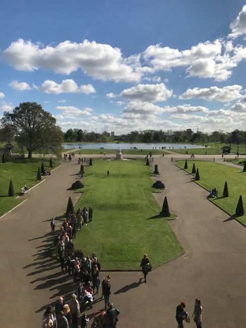 A detailed view of a well-maintained landscaped area in Kensington Gardens featuring a prominent memorial monument with a tall, ornate spire and sculptures. Surrounding the monument are lush green tre