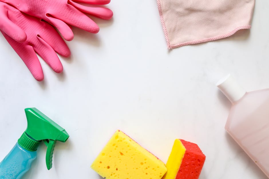 Arrangement of cleaning supplies on a white surface, including a pair of pink rubber gloves, a pink cloth, a green and blue spray bottle, a yellow and red sponge, and a plastic white bottle, all used for surface cleaning and sanitisation in residential or commercial environments, as part of domestic cleaning services offered by [COMPANY_NAME] for end of tenancy cleaning in Earls Court flats, West Kensington.