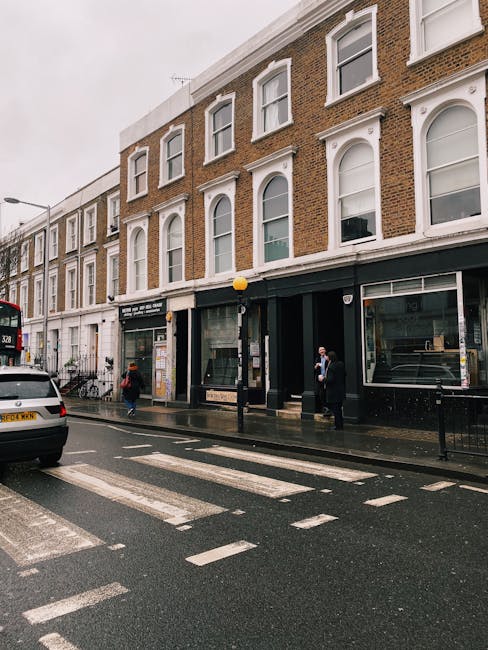 Street view of a three-story brick building with large windows and white decorative trims, situated on a wet urban road with a pedestrian crossing and parked cars. The ground floor features commercial spaces with large glass storefronts and black frames, some displaying interior furniture or items. A few pedestrians are seen walking on the sidewalk, one wearing a face mask. Overhead street lighting illuminates the scene on a cloudy day, highlighting the damp pavement and reflective surfaces. This image exemplifies the importance of surface cleaning and regular maintenance for commercial premises, as highlighted by [COMPANY_NAME] in providing deep cleaning and sanitisation services for urban environments, especially during periods of late access or delays that can impact cleanliness and hygiene standards.