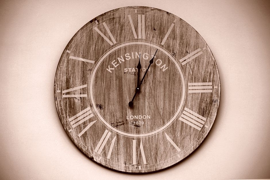 A large round wall clock with a rustic wooden finish and white Roman numerals, displaying a time just past 12:00. The clock has the text 'Kensington Station' and 'London 1809' printed on its face. It is mounted on a light-colored wall, with soft ambient lighting highlighting the surface details. The clock's surface appears clean, though the wood grain and numerals are clearly visible, emphasizing its vintage style. This clock is situated in an interior space, potentially a living room or office, and serves as a decorative yet functional timepiece. The focus on surface detail and material texture reflects a high standard of interior decor, consistent with professional surface and deep cleaning practices that keep such items free from dust and grime, similar to the services provided by [COMPANY_NAME] in the context of domestic and commercial cleaning. Also, it exemplifies the importance of proper maintenance for interior furnishings to ensure hygiene and aesthetic appeal.