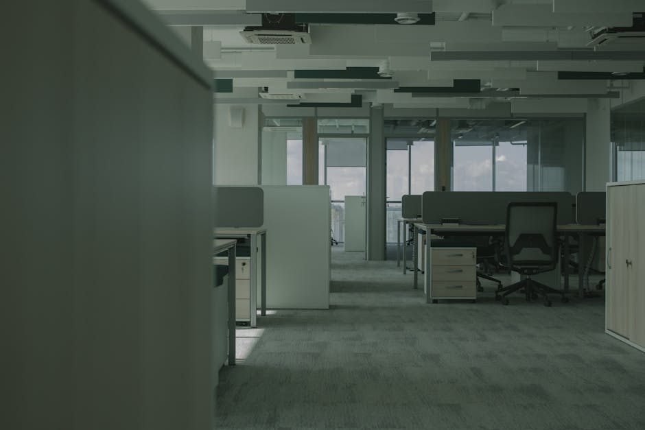 Interior view of a modern office space with several workstations equipped with desks, chairs, and partition panels. The carpeted floor appears clean and well-maintained, with natural light filtering through large windows at the far end. The ceiling features a suspended design with integrated lighting and ventilation systems. The space is tidy, with no visible dust or clutter, reflecting professional cleaning standards. The overall environment conveys a sense of hygiene and freshness, ideal for business operations. The image represents a typical commercial setting that could benefit from deep cleaning and surface sanitisation, as offered by [COMPANY_NAME] for office carpet cleaning in Kensington.