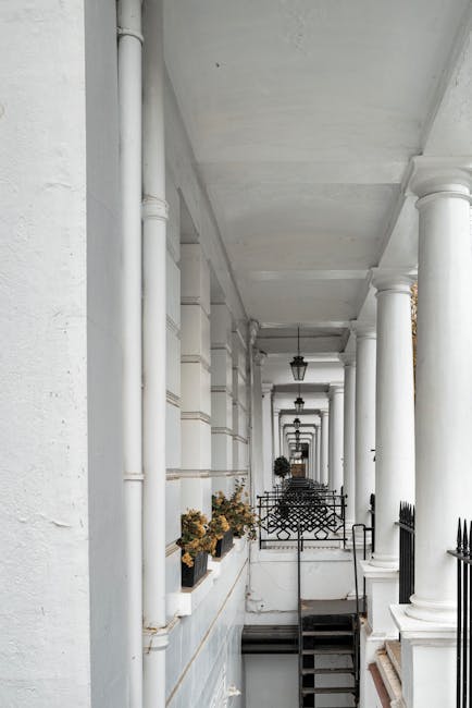 A white-painted residential exterior corridor with a series of tall, cylindrical columns supporting the ceiling, creating a colonnade effect. The corridor features decorative black wrought iron fencing along the edge and is lined with potted plants containing dried flowers. The surfaces, including walls, columns, and ceiling, appear clean and well-maintained under natural daylight. The floors are not visible, but the overall scene emphasizes a neat, tidy appearance suitable for surface cleaning or maintenance in a domestic setting. The image reflects a focus on architectural cleanliness and presentation, which is relevant to professional cleaning services such as those offered by carpetcleanerswestkensington.co.uk in the context of domestic cleaning and surface sanitisation.