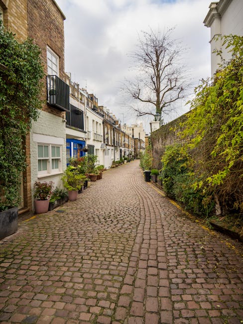 A narrow residential street in West Kensington W14, featuring a cobblestone surface with a slight curve. On the left side, there are multi-story houses with white facades, some with balconies and black railings, and window planters filled with plants. On the right side, a stone wall covered partially with green foliage runs alongside the street. A tall, leafless tree stands in the background against an overcast sky, casting diffused light over the scene. The street appears clean and well-maintained, with no visible debris or litter, reflecting a tidy and quiet neighborhood typical of West Kensington. This image illustrates an exterior urban environment suitable for demonstrating surface cleaning or maintenance services offered by companies like [COMPANY_NAME], aligned with the focus on the real cost of carpet cleaning in West Kensington W14.