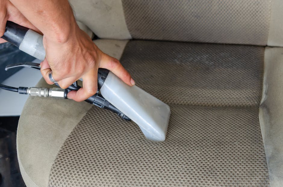 A hand holding a handheld upholstery cleaning machine is visibly working on a beige fabric sofa seat with a textured, patterned cushion. The machine has a gray base and a black handle, with its nozzle pressed against the surface to perform deep cleaning and sanitisation. The surrounding area shows other sections of the sofa in a similar light beige color, with natural lighting illuminating the room, highlighting the clean surface and the focused cleaning process by [COMPANY_NAME], which specializes in domestic cleaning services. The room appears tidy, with an emphasis on maintaining hygiene and surface cleanliness of upholstery furniture.