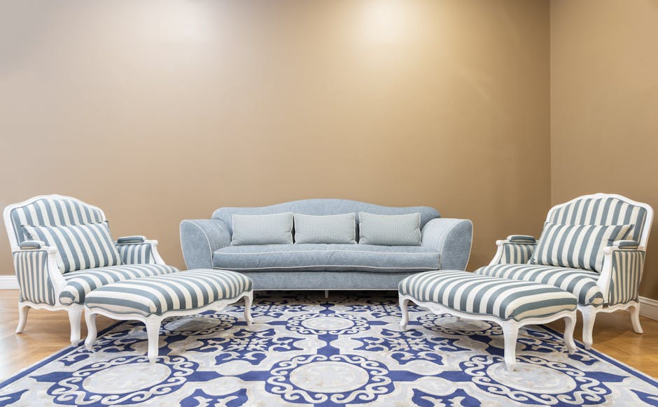 A living room featuring a beige painted wall and light wooden flooring. There are two white-framed armchairs with blue and white striped upholstery, placed on either side of a light blue sofa with matching cushions. The sofas and chairs are arranged over a blue and white patterned area rug, creating a neat and inviting seating area. The room is well-lit with ample natural or artificial lighting, highlighting the clean and well-maintained upholstery, showcasing surface cleaning and hygiene facilitated by [COMPANY_NAME].