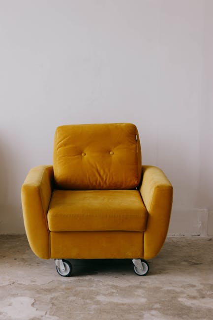 A hand holding a handheld upholstery cleaning machine is visibly working on a beige fabric sofa seat with a textured, patterned cushion. The machine has a gray base and a black handle, with its nozzle pressed against the surface to perform deep cleaning and sanitisation. The surrounding area shows other sections of the sofa in a similar light beige color, with natural lighting illuminating the room, highlighting the clean surface and the focused cleaning process by [COMPANY_NAME], which specializes in domestic cleaning services. The room appears tidy, with an emphasis on maintaining hygiene and surface cleanliness of upholstery furniture.